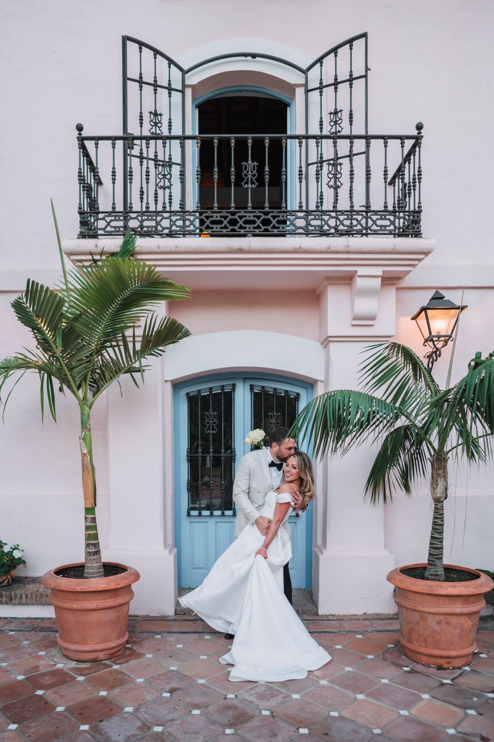 A romantic wedding photo in front of a pink Mediterranean building with ornate balcony and palm trees.