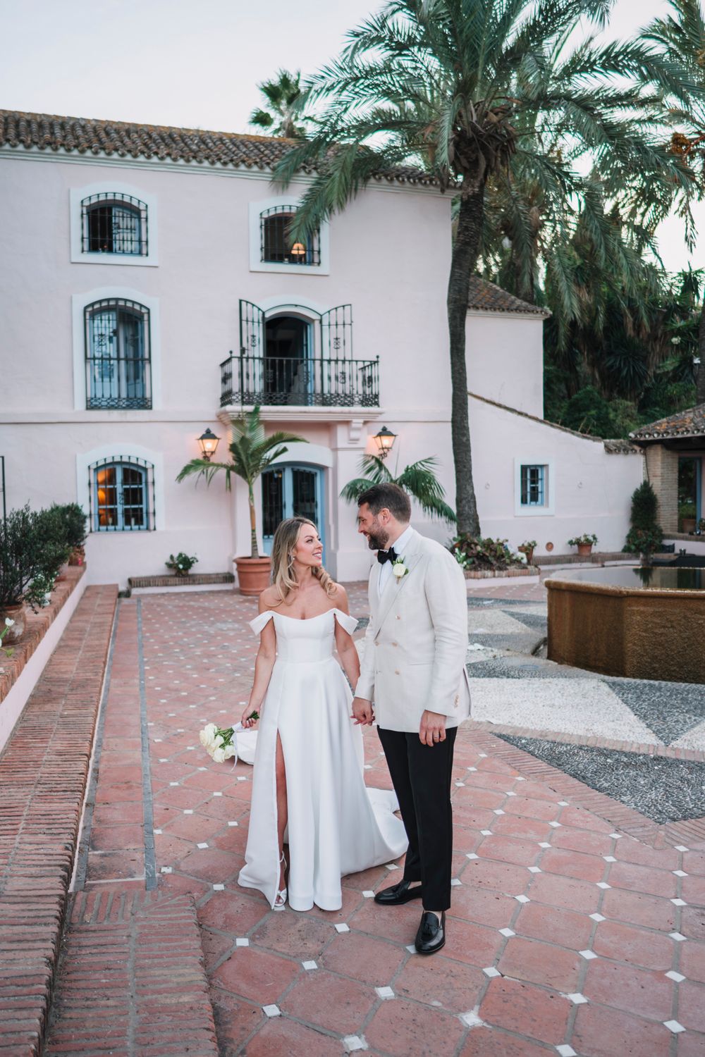A wedding couple poses in front of a Mediterranean-style villa with blue windows and palm trees.