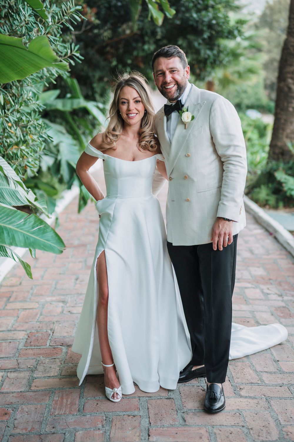 A joyful couple in elegant white wedding attire poses on a brick pathway surrounded by greenery.