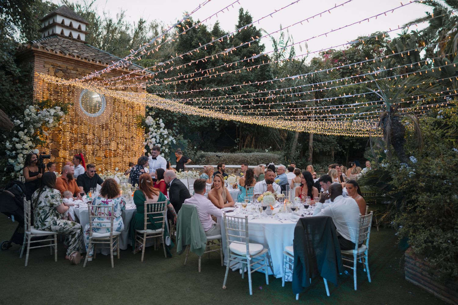 An outdoor wedding reception under twinkling string lights with guests seated at white tables in a garden setting.