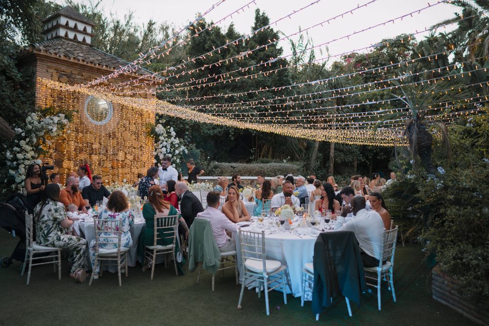 An outdoor wedding reception under twinkling string lights with guests seated at white tables in a garden setting.