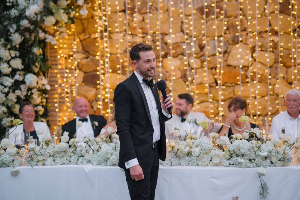 Wedding reception head table decorated with white floral arrangements and illuminated by warm string lights.