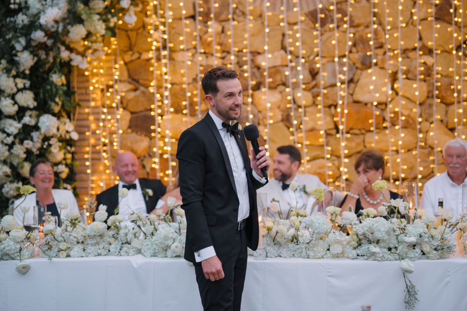 Wedding reception head table decorated with white floral arrangements and illuminated by warm string lights.