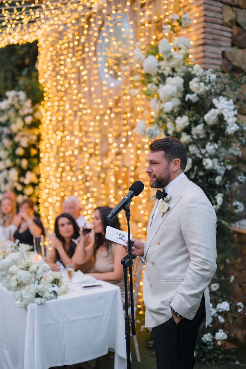 A speaker gives a wedding speech at an elegantly decorated outdoor reception with fairy lights.