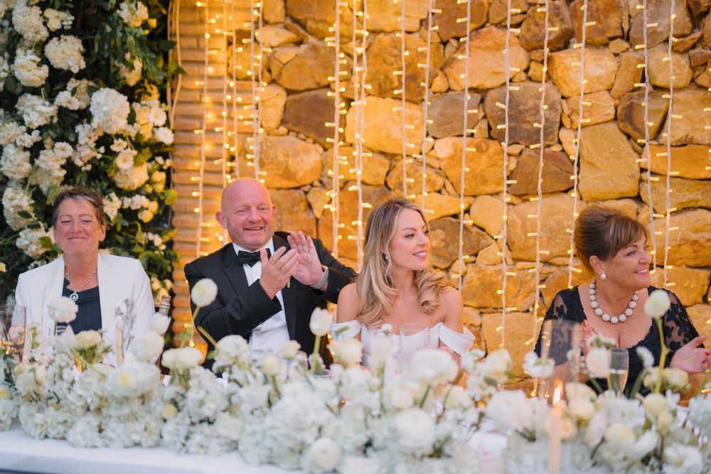 Wedding party seated at head table with white floral decorations and glowing string light backdrop.