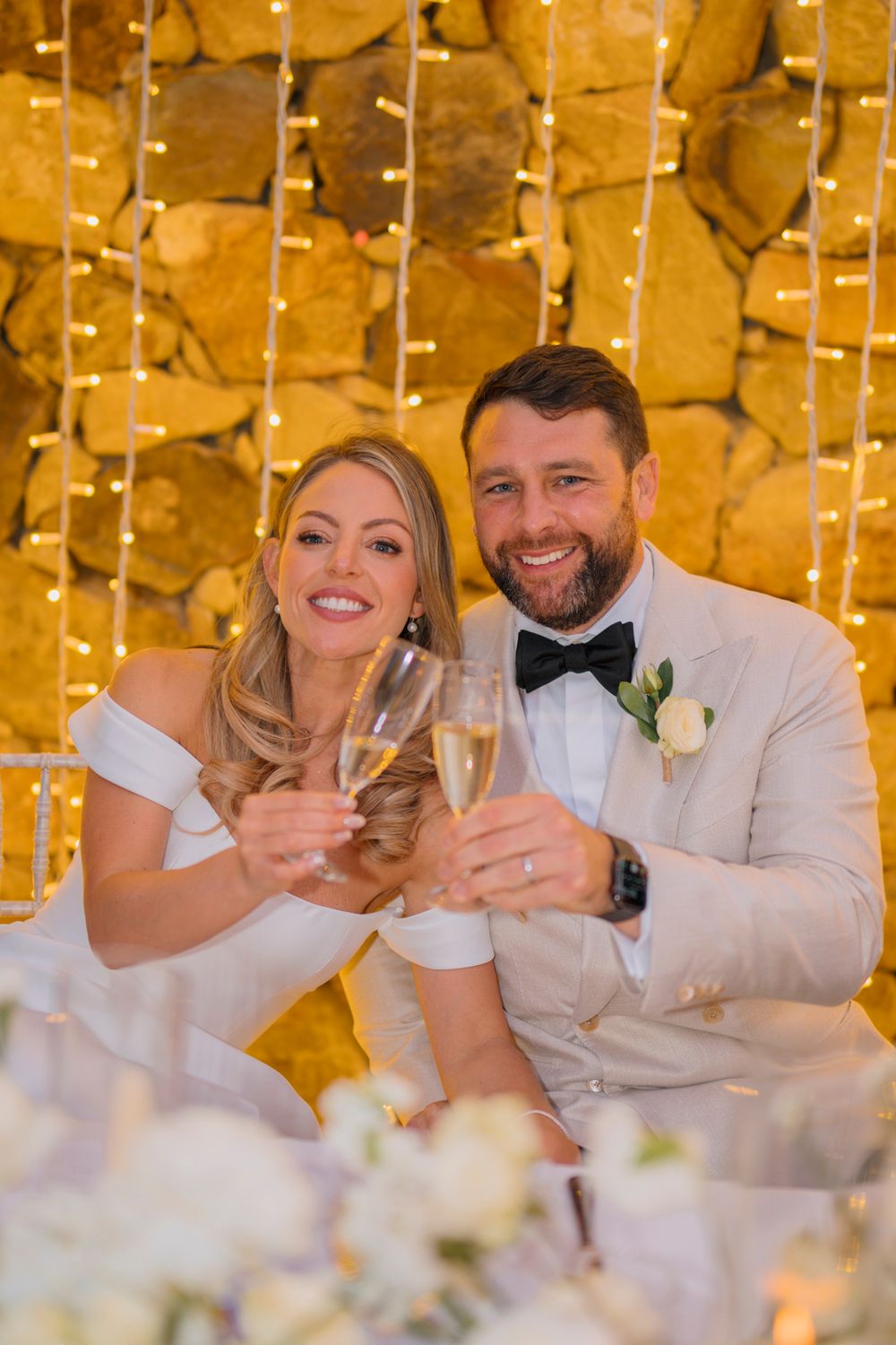 Newlyweds toast with champagne glasses against a backdrop of golden fairy lights and stone wall.
