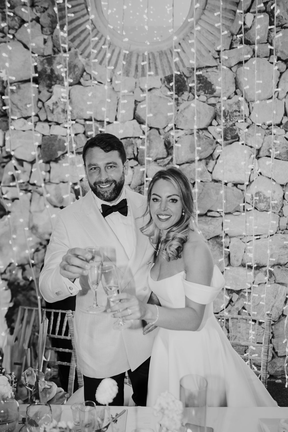 Black and white portrait of newlyweds toasting with champagne against a stone wall and fairy lights.