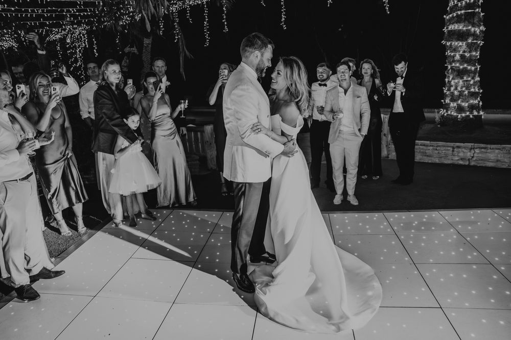 Black and white capture of a wedding first dance surrounded by guests on a lit dance floor.