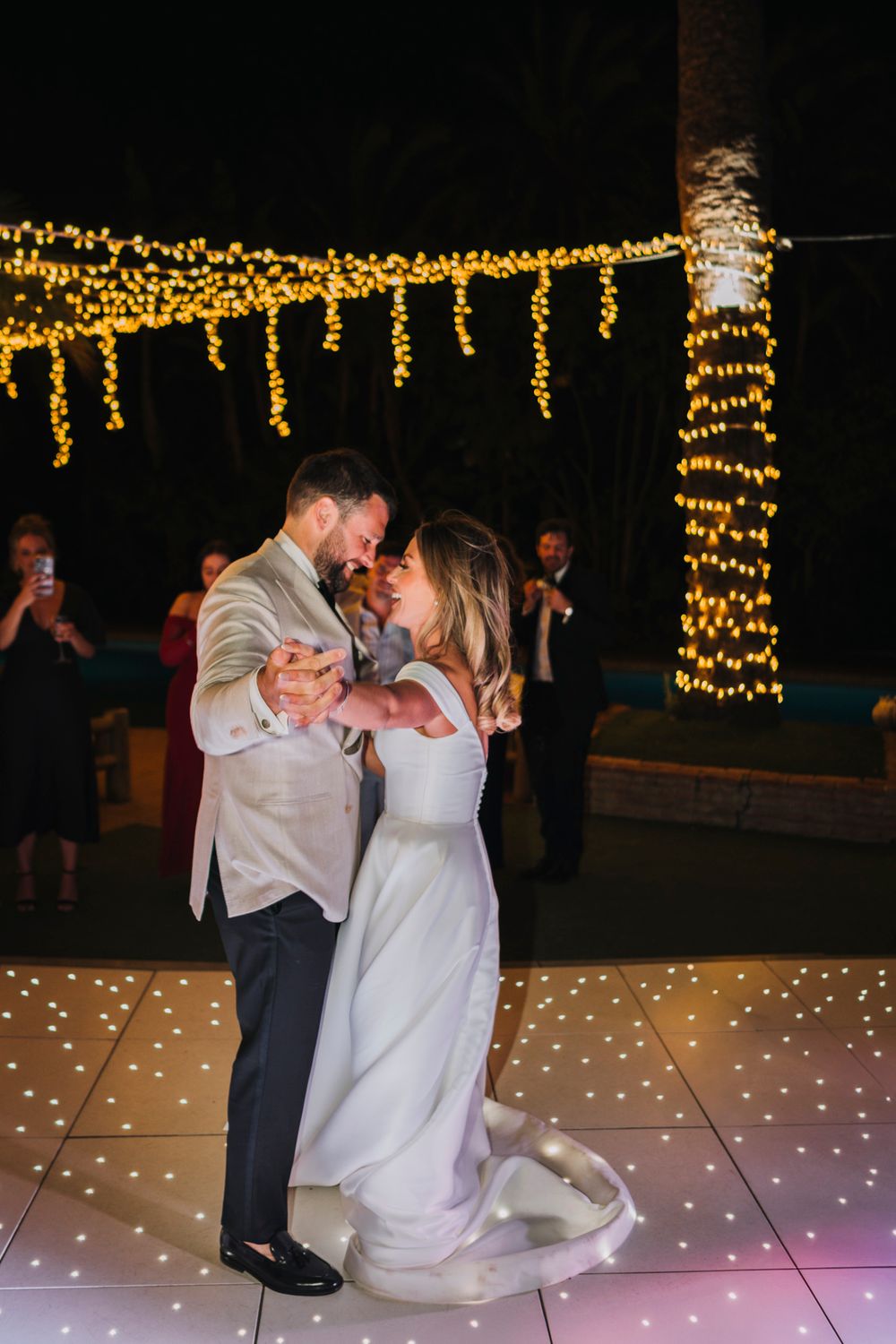 Couple shares their first dance on an illuminated dance floor under golden string lights.