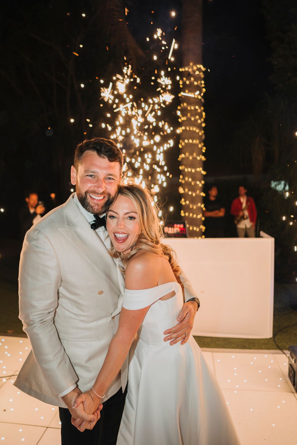 Newlyweds pose together against a backdrop of sparkling fireworks and fairy lights.