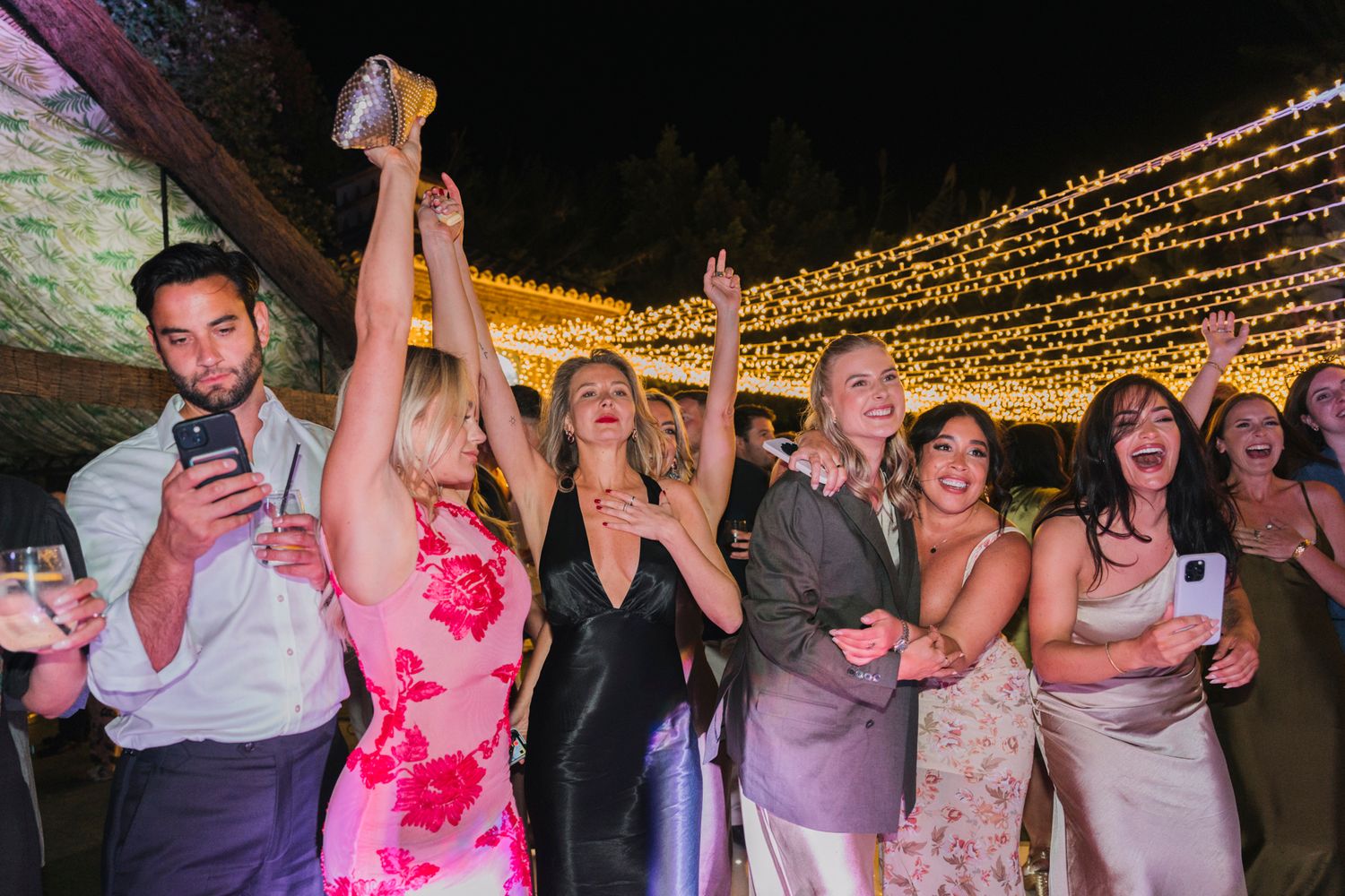 Wedding guests celebrate joyfully under twinkling string lights at an outdoor evening reception.