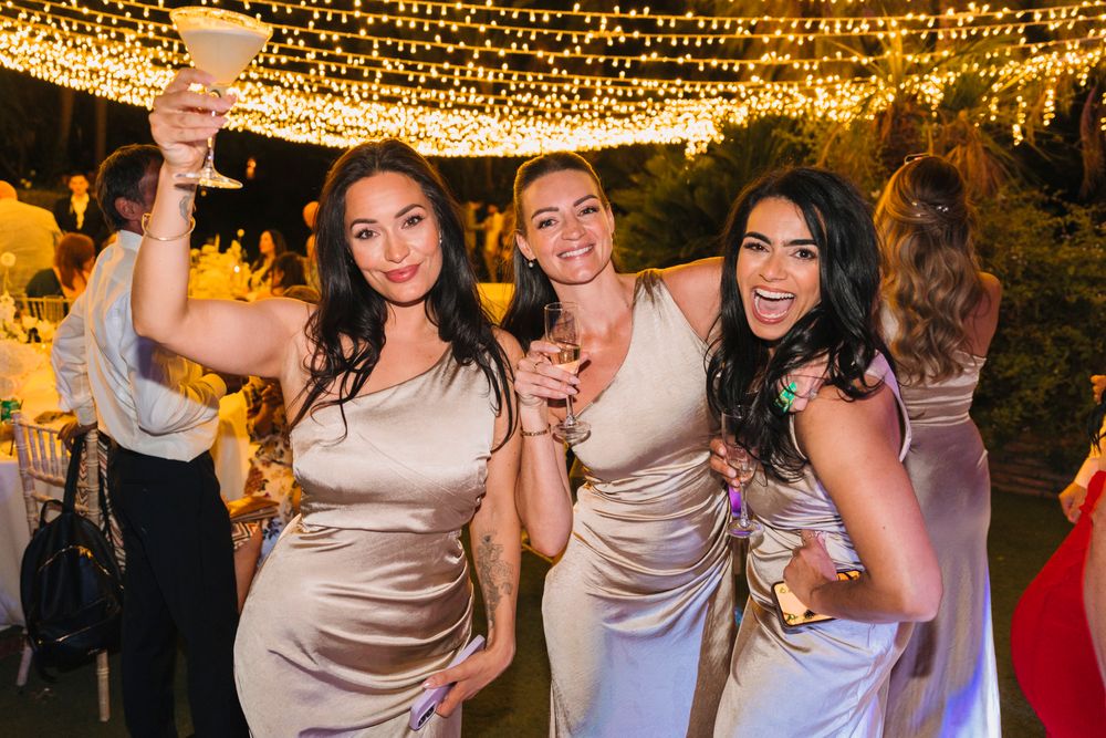 Three attendees in champagne-colored dresses pose with drinks under glowing string lights at an evening celebration.