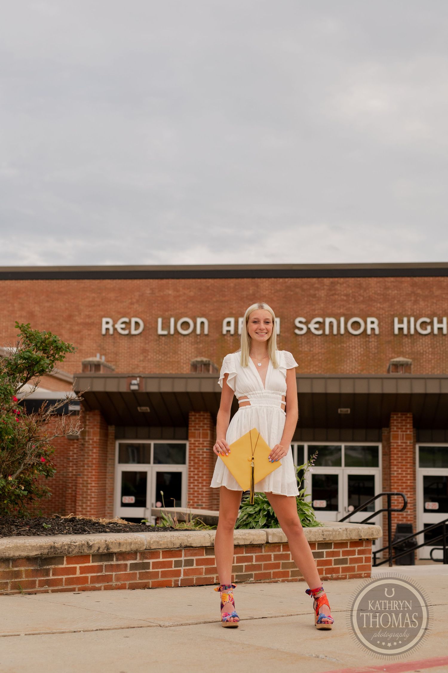 Graduate poses confidently in white dress and yellow graduation attire outside high school entrance.