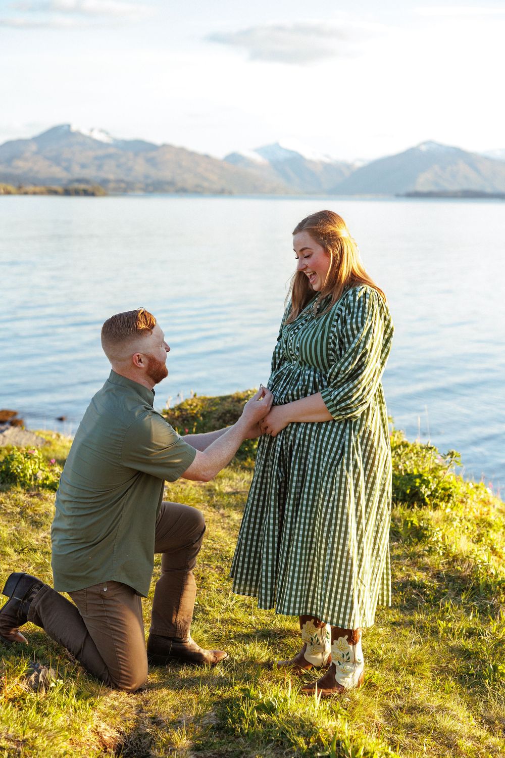 A couple sharing a romantic proposal moment on a grassy hillside overlooking mountains and water during golden hour.