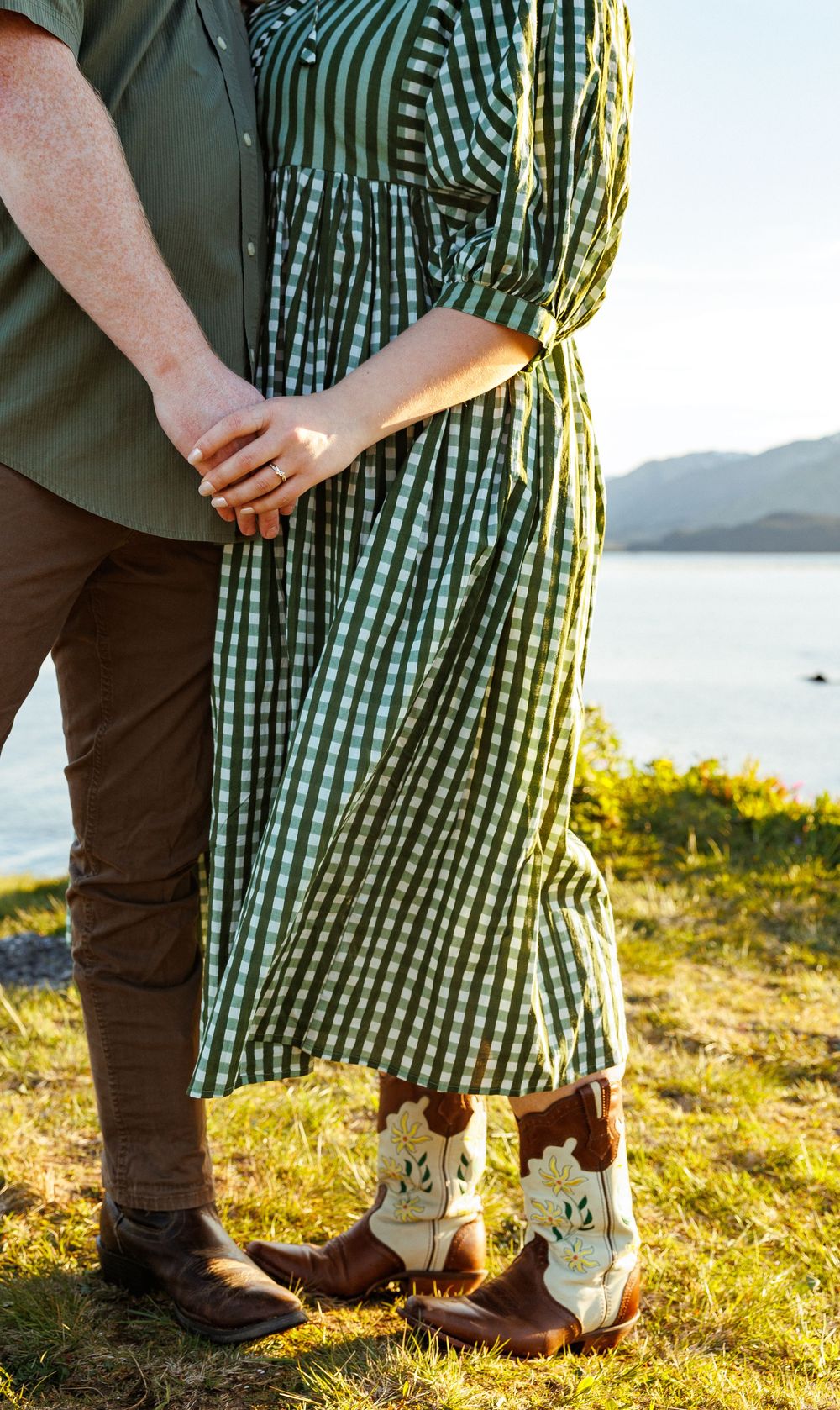 Couple in gingham dress and brown pants hold hands by lake wearing matching cowboy boots at sunset.