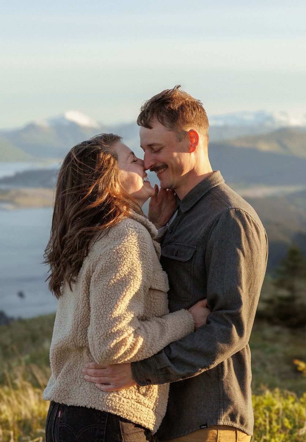A romantic couple embraces during golden hour on a mountaintop overlooking a scenic coastline.