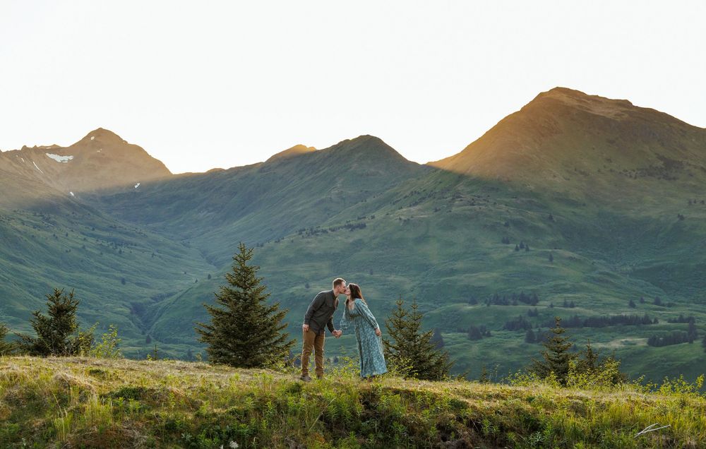 A scenic mountain landscape with green grass and pine trees against rugged peaks at golden hour.