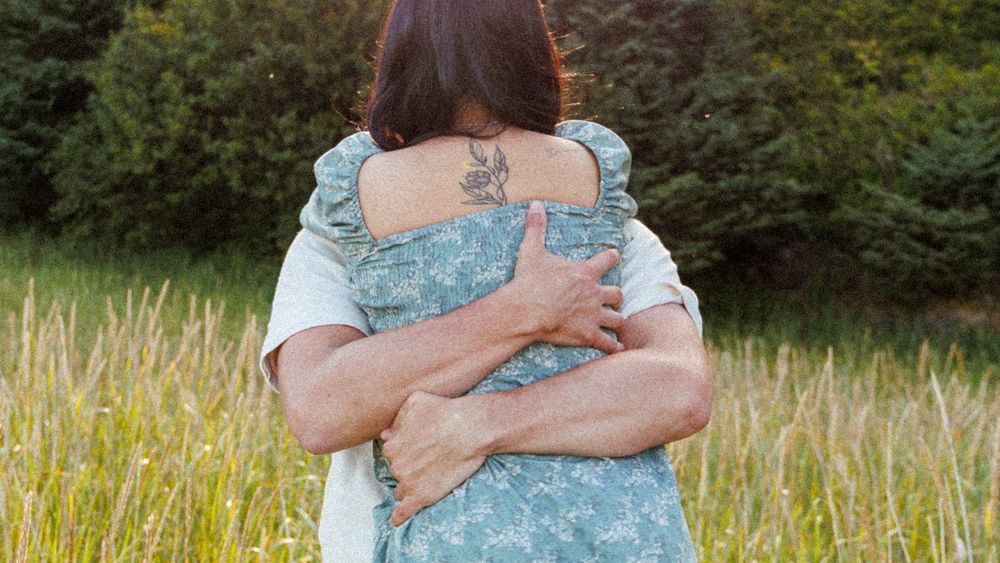 Someone wearing denim overalls hugs another person outdoors in a field of tall grass at sunset.
