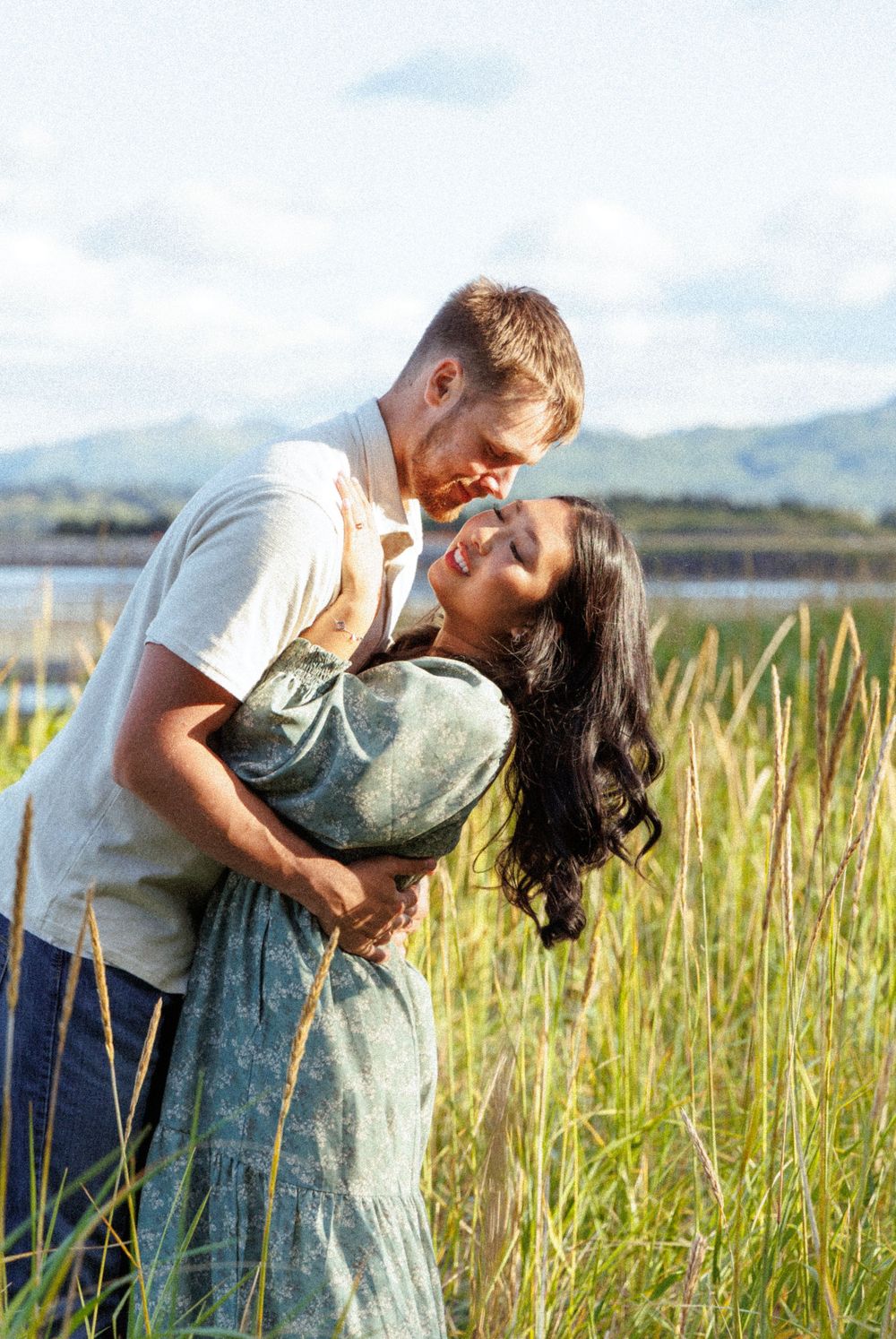 A couple shares a romantic moment in a tall grass field on a sunny day, creating a dreamy outdoor scene.
