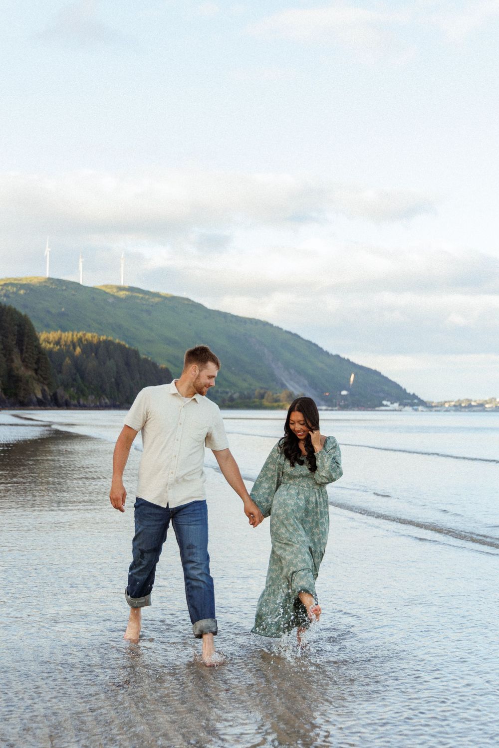 A couple walks hand in hand along a misty beach shoreline with coastal mountains in the background.