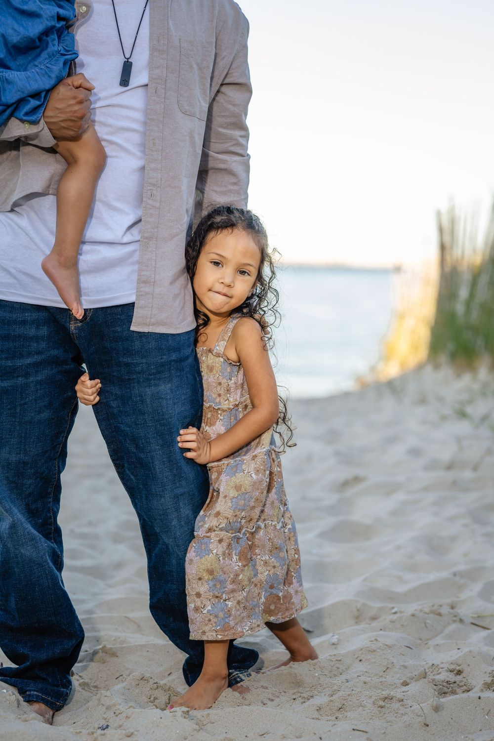 A young child in a floral dress hugs their parent's leg while standing barefoot on the beach.