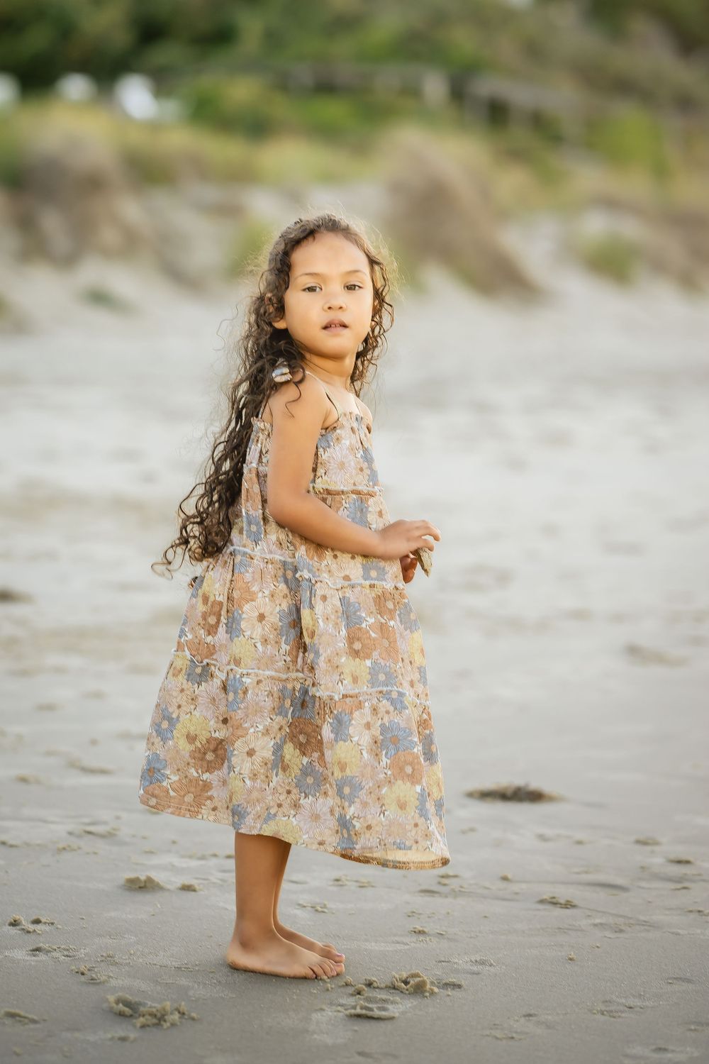 A young child in a floral dress stands barefoot on a sandy beach.