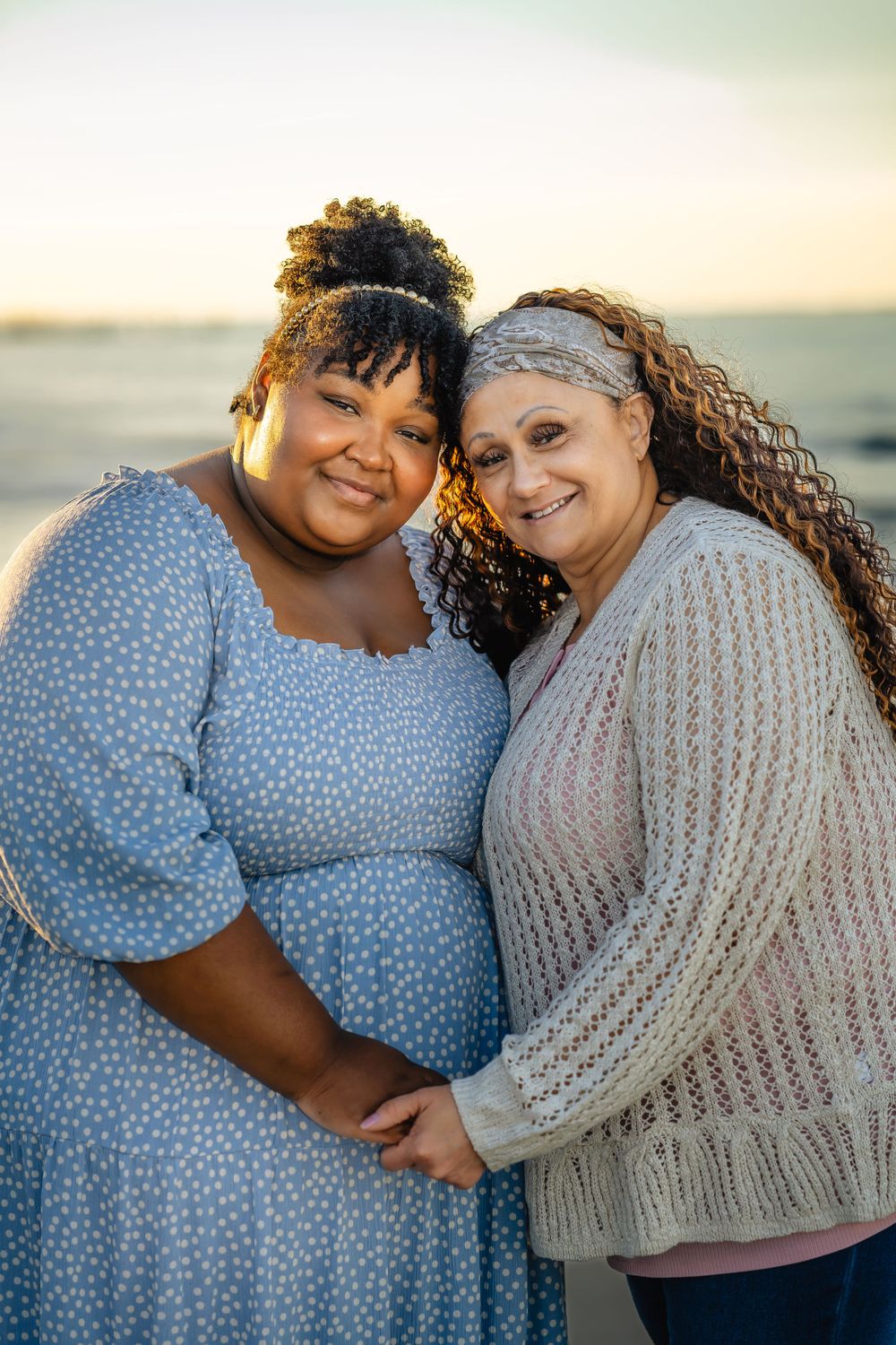 Mother and daughter wearing blue and beige sweaters embrace on the beach at golden hour.