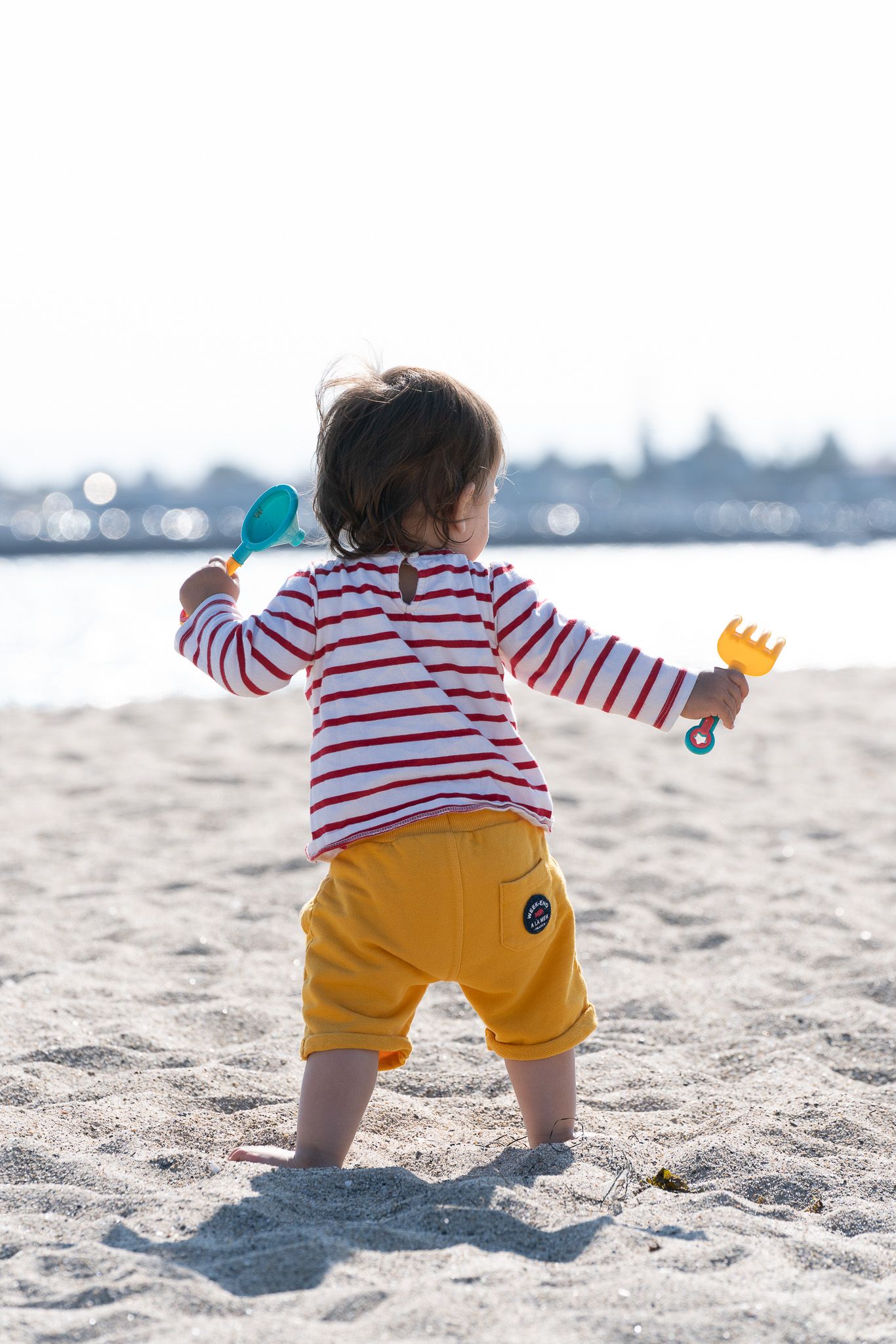 Toddler in striped shirt and yellow shorts plays with beach toys on sandy shore.  Located near the Santa Cruz Wharf, California.