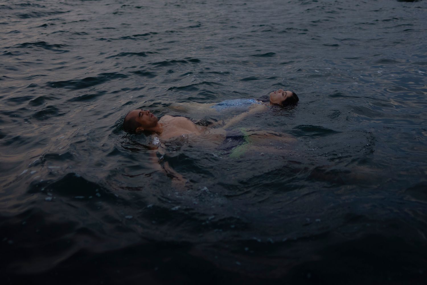 A couple floats in the dark lake water. They float on their back, facing opposite directions, and hold hands in Lake Ontario, Kingston, CA