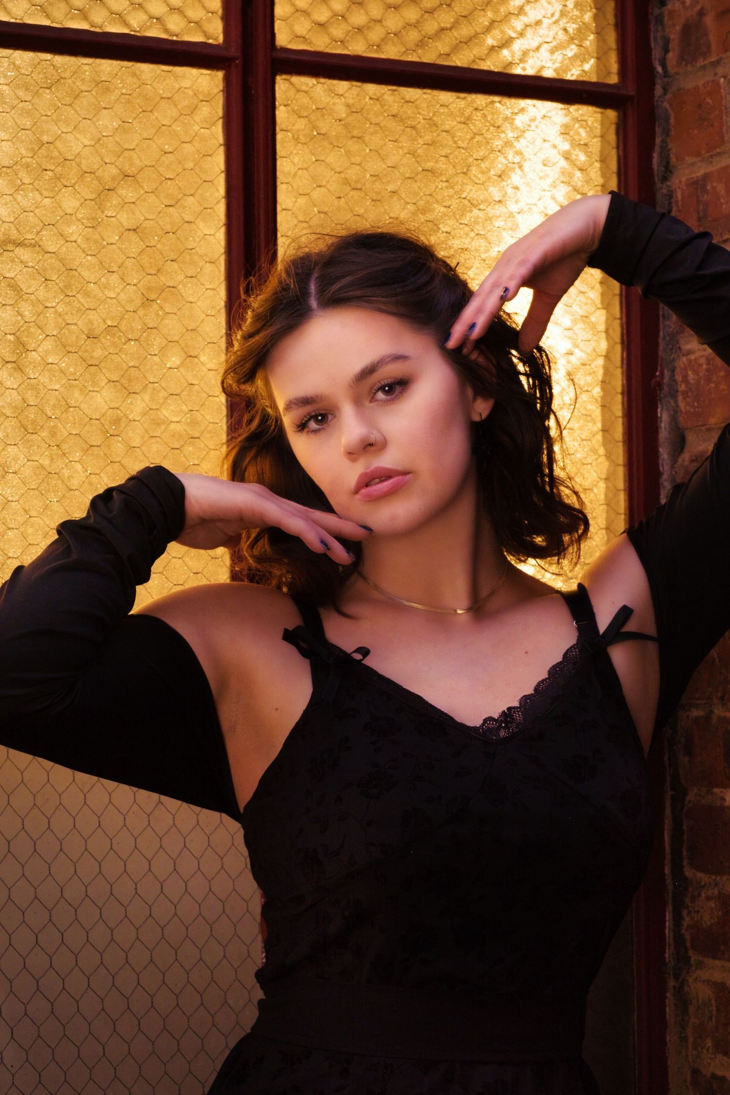 Moody portrait in black lace dress against golden backlit window with dramatic lighting.