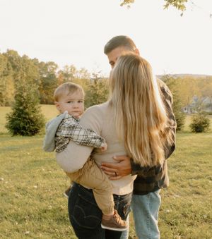 Young family embracing in a sunny meadow with evergreen trees in the background at golden hour.