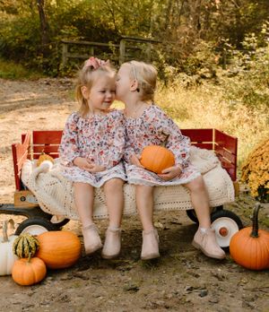 Two children in floral dresses share secrets on red wagon surrounded by fall pumpkins.