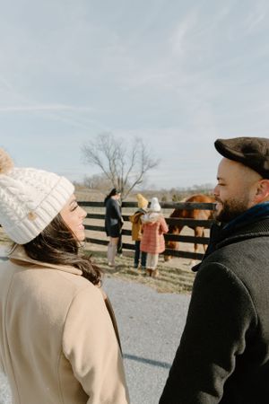 A winter scene with two people chatting by a horse fence wearing warm knit hats.