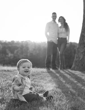 Black and white portrait of a small child sitting in grass with two adults in background.