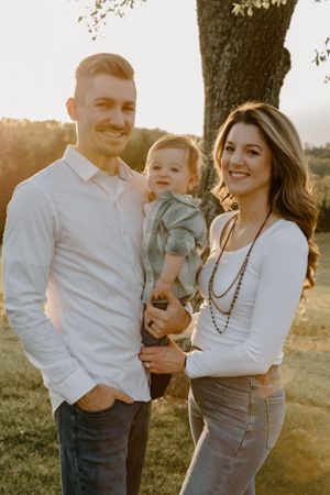 A young family poses together outdoors during sunset against a tree backdrop.