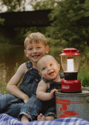 Children sit on picnic blanket with red camping lantern by peaceful water.