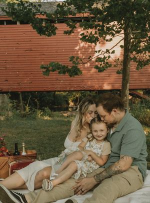 Family having picnic under tree against rustic red building at sunset.