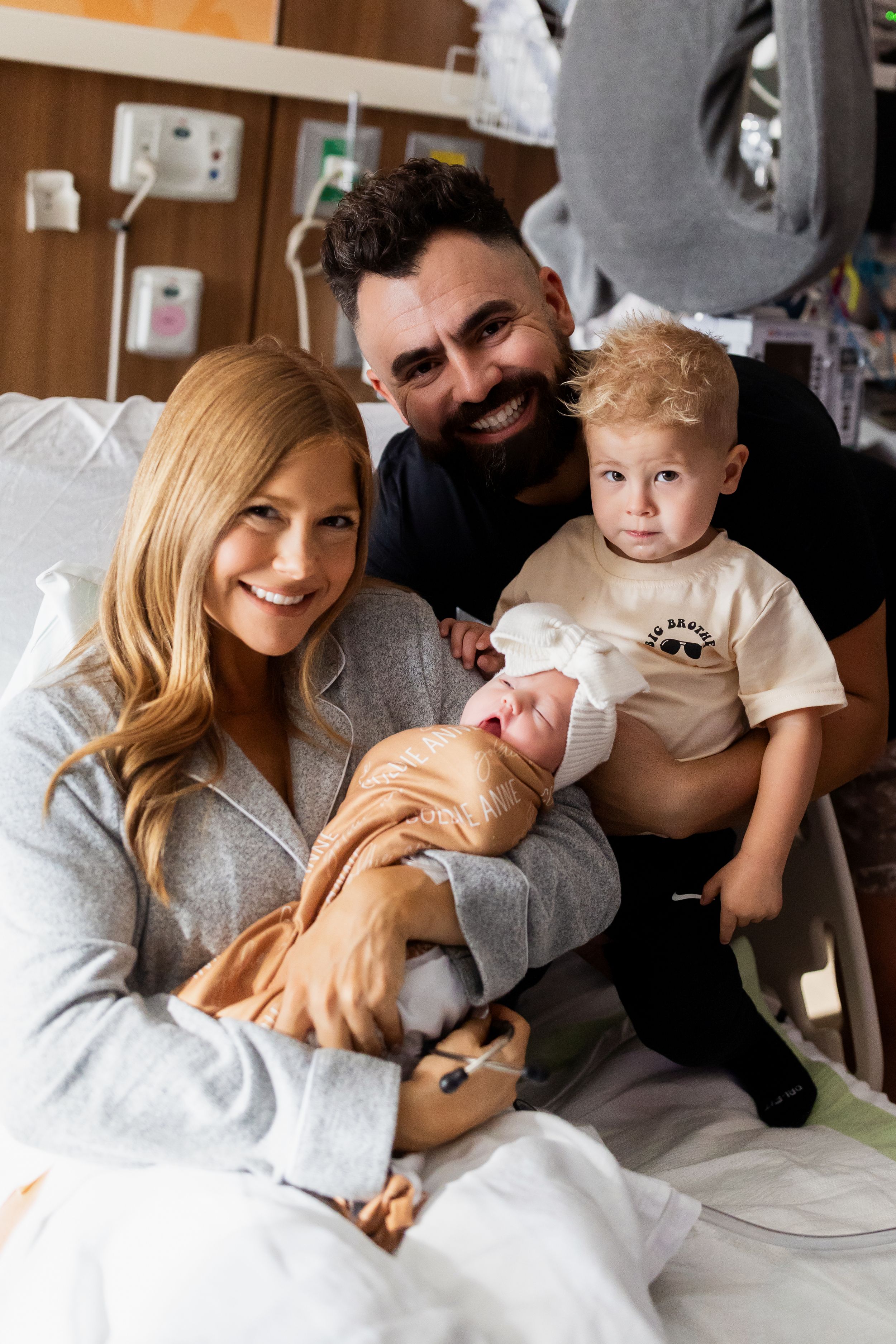 A family cuddles together in a hospital room, with a newborn baby and toddler joining their tender moment.
