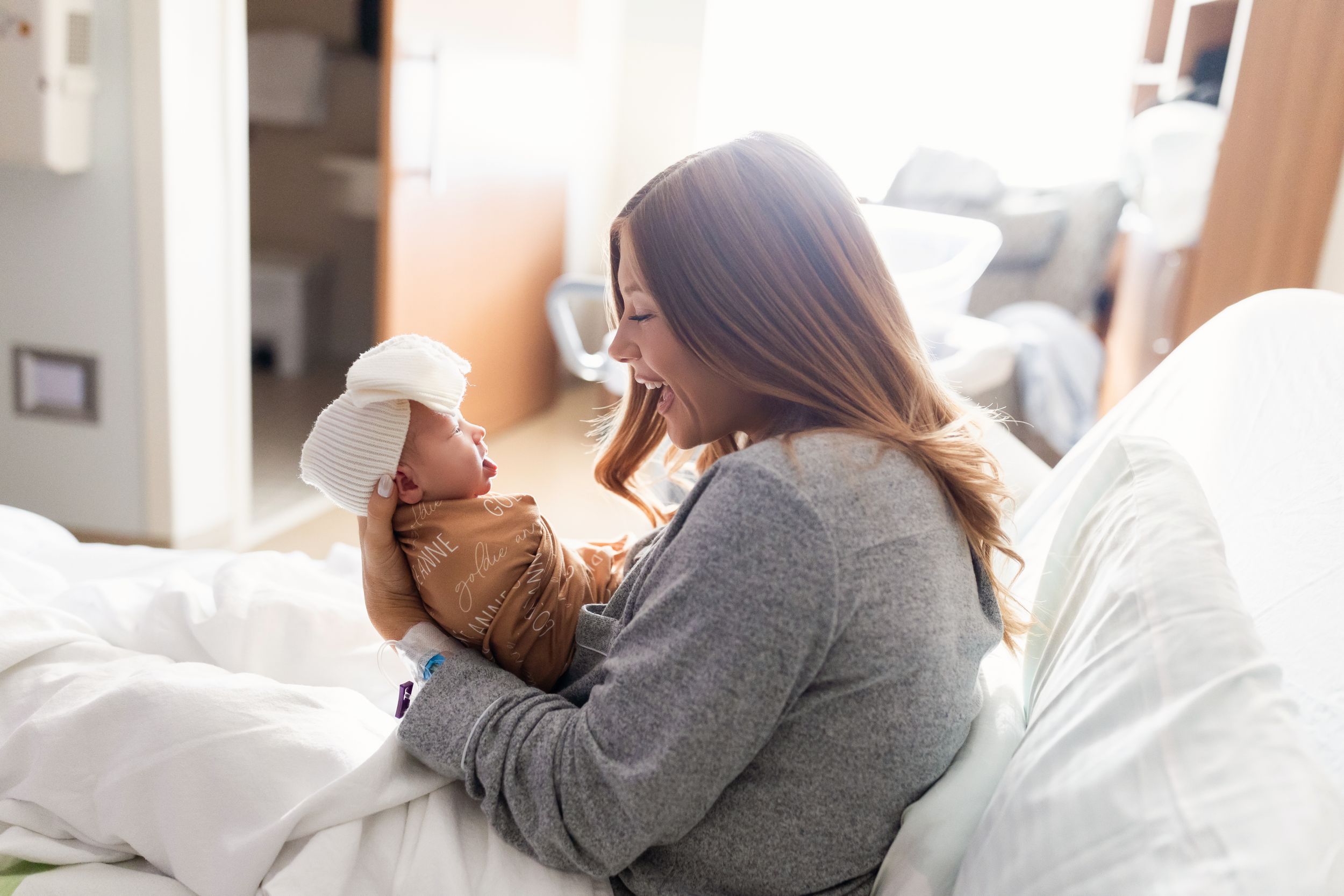 A parent in a gray sweater tenderly holds and gazes at their newborn baby while sitting on a white bed.