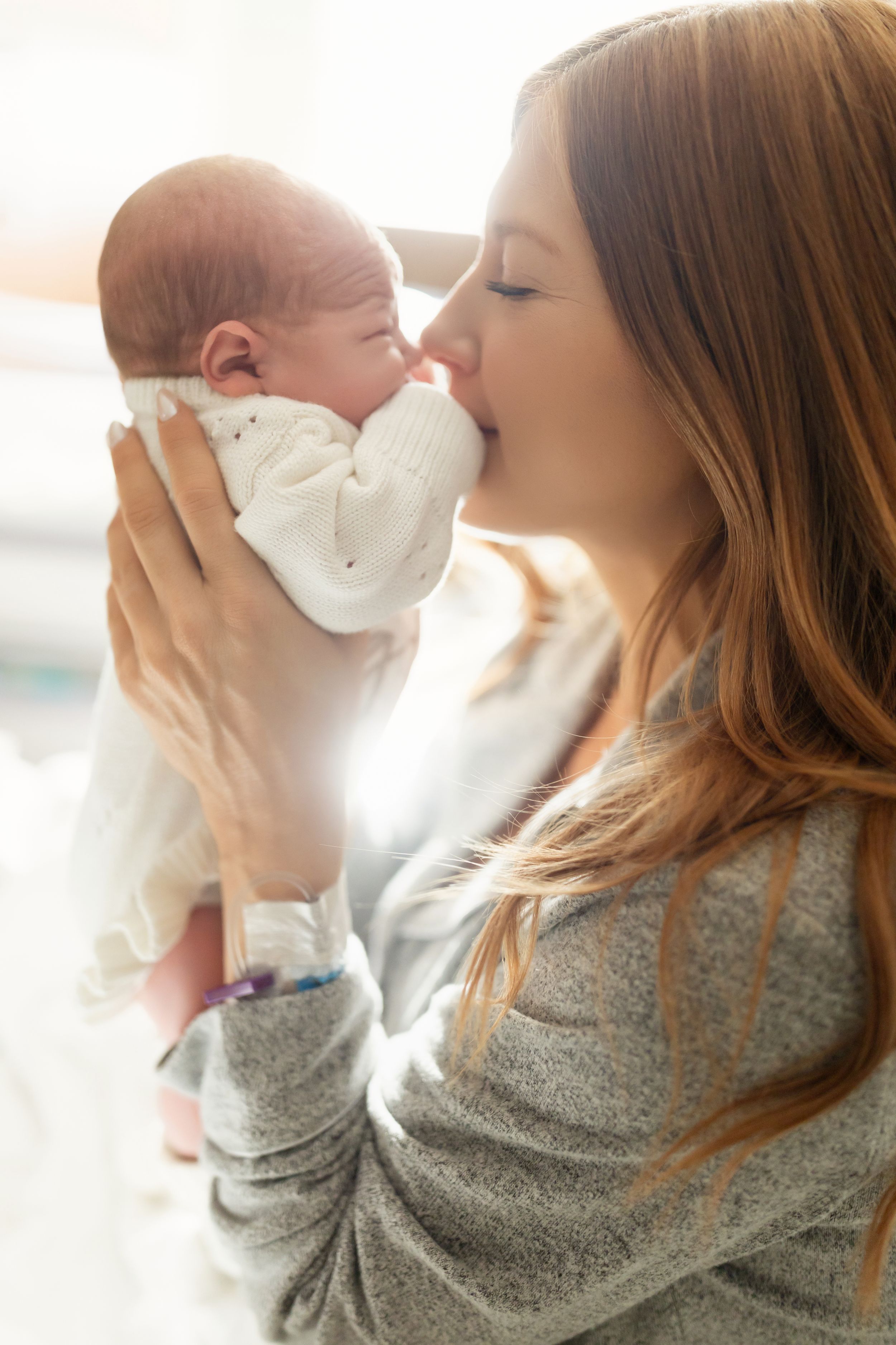 Woman in gray sweater holding and kissing newborn baby in white blanket in bright sunlit room.