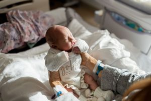 Newborn baby wearing white onesie rests on hospital bed while being cared for by medical staff.