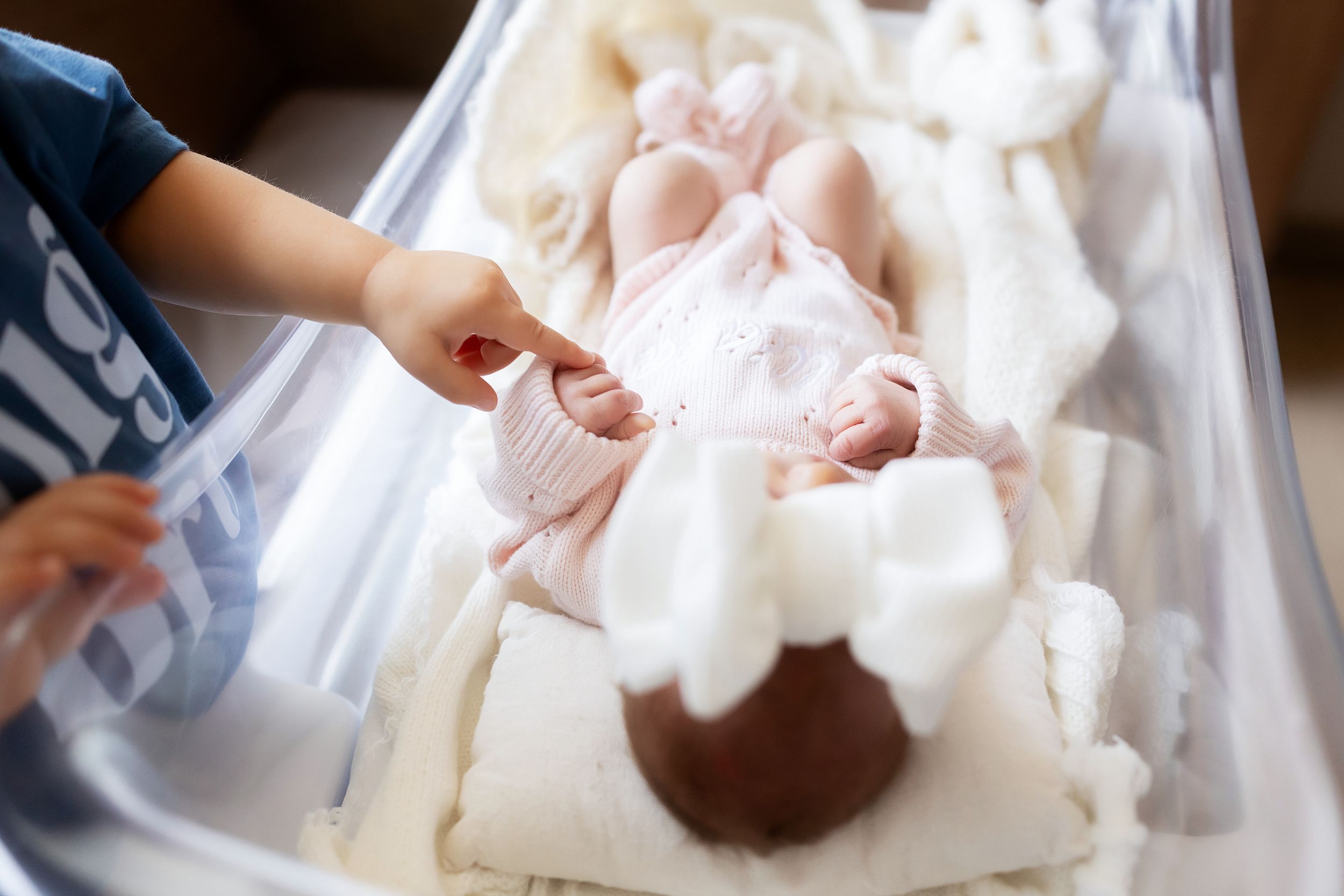 Newborn baby in hospital bassinet wearing pink outfit, with sibling's hand gently reaching to touch them.