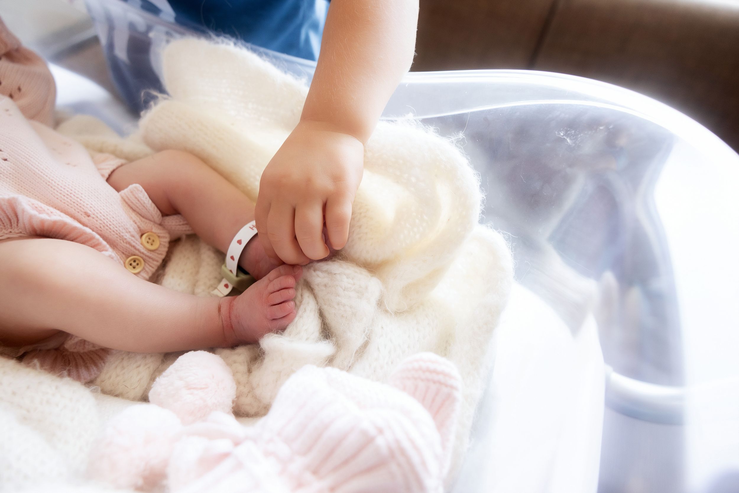 Toddler gently holding newborn baby's hand on a soft white blanket in a bassinet.