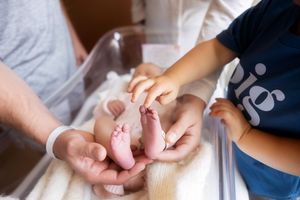 Adult hands gently hold tiny newborn baby feet while a young child touches them in a hospital nursery.