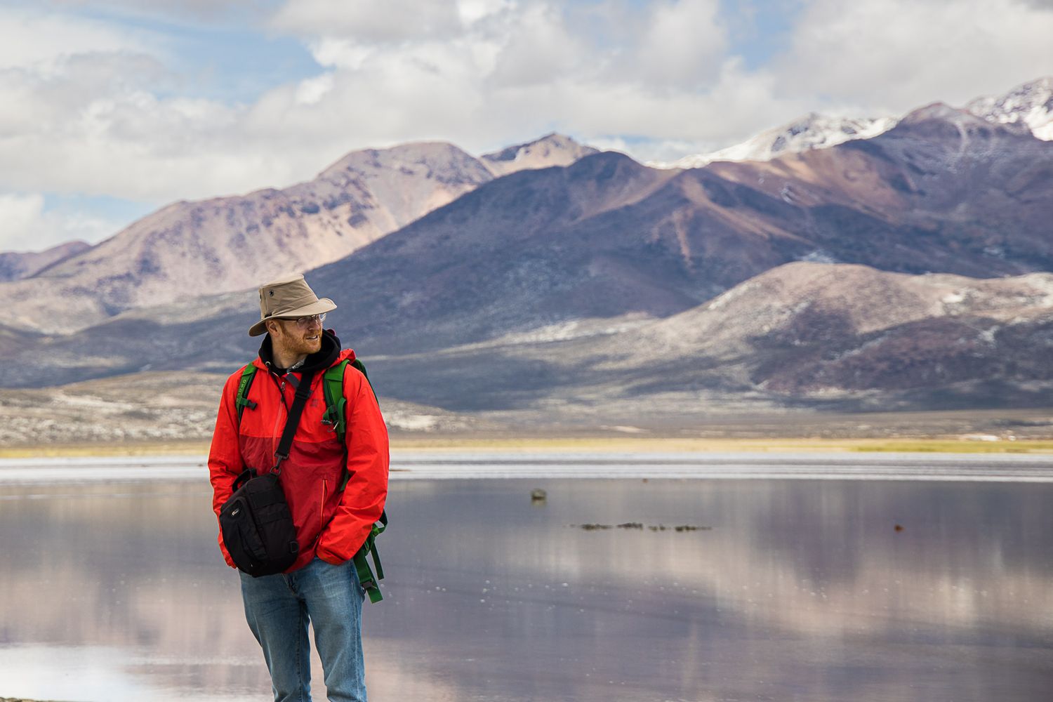 Photographe vêtu d'un manteau rouge explorant le paysage montagneux accidenté qui borde un lac alpin serein, au Pérou.