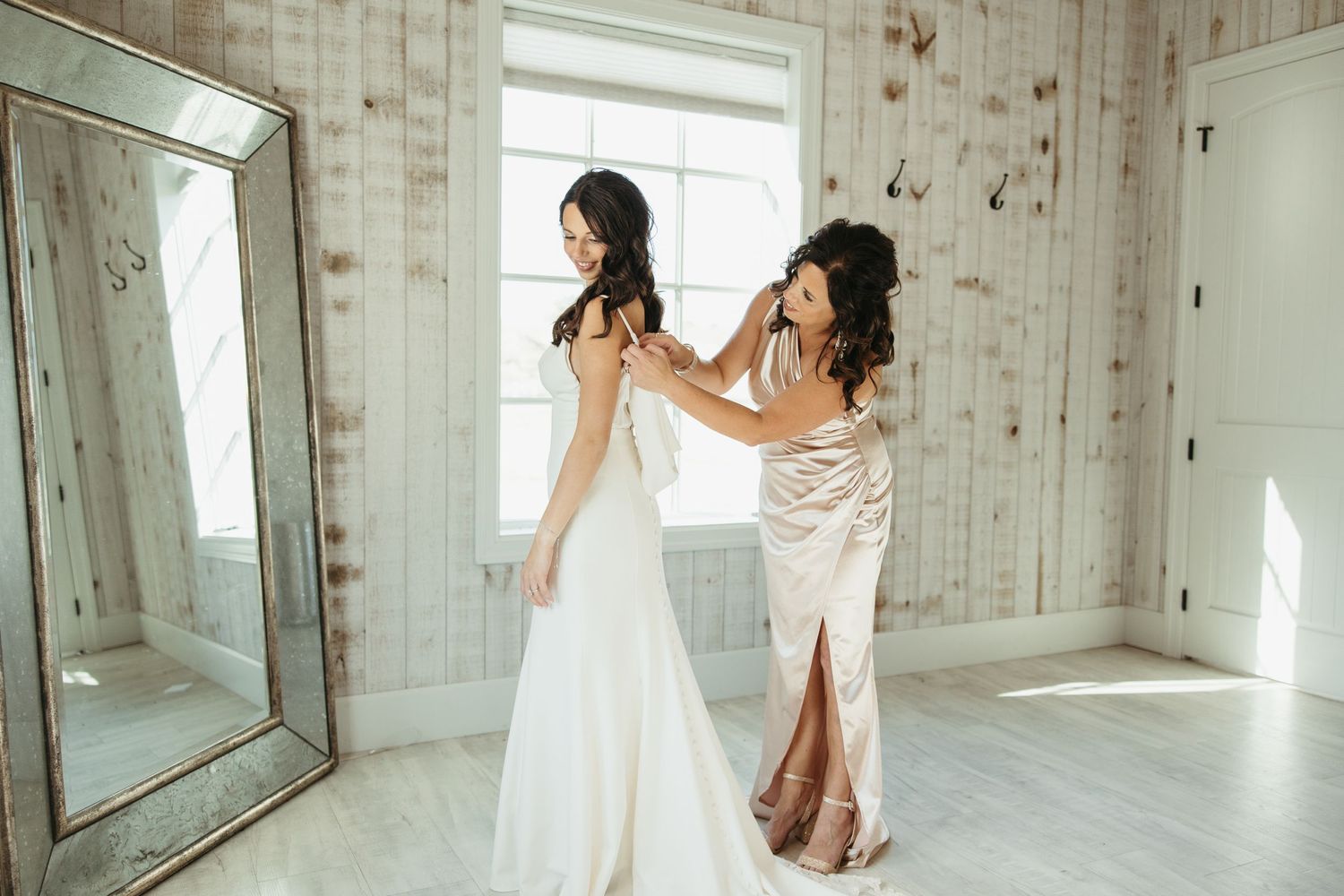 Wedding preparation scene showing final dress adjustments in a rustic room with mirrors.