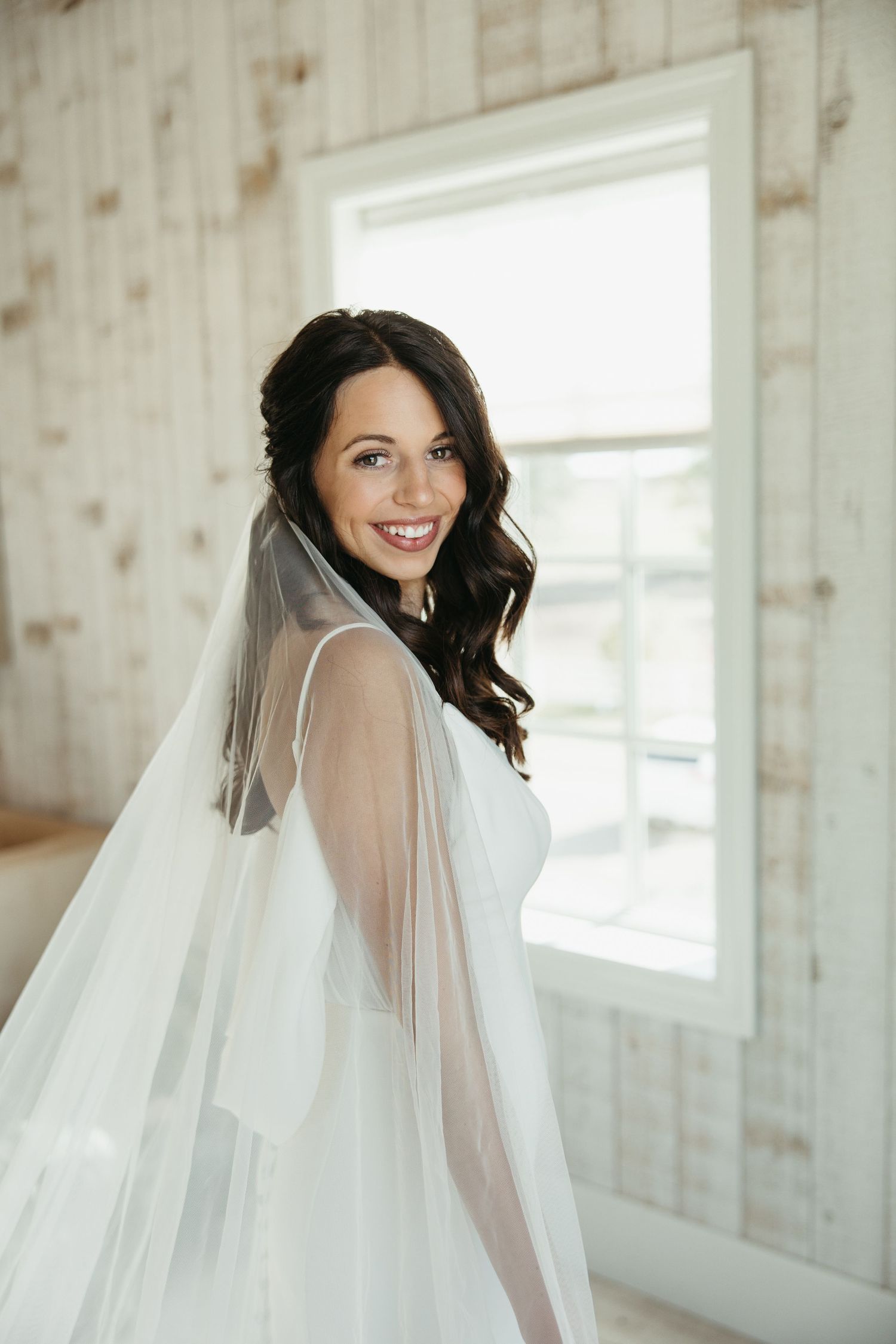 Side profile of someone in a white wedding dress and veil smiling near a window.