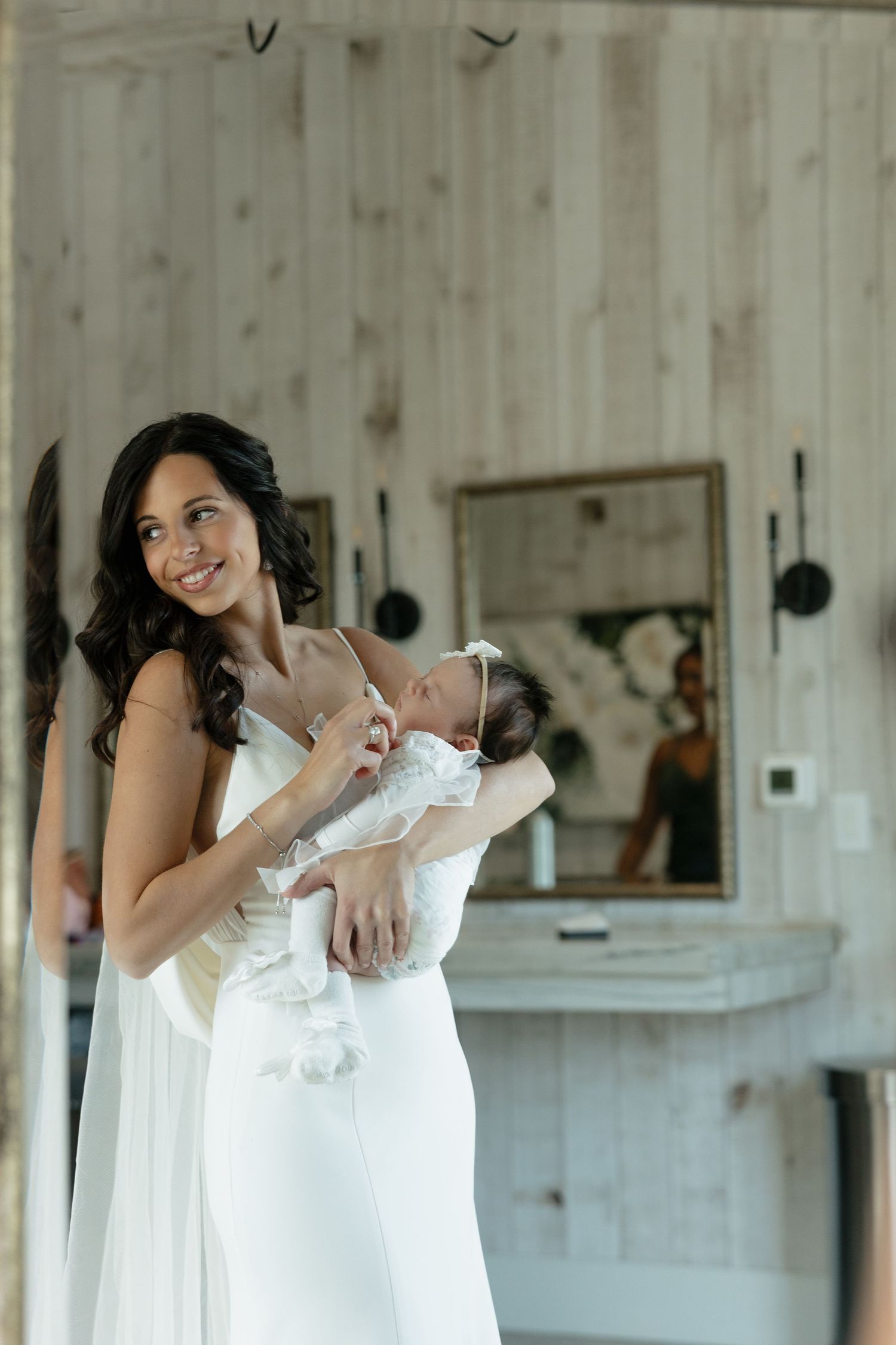 A mother in an elegant white wedding dress holds her baby in a rustic wooden room.