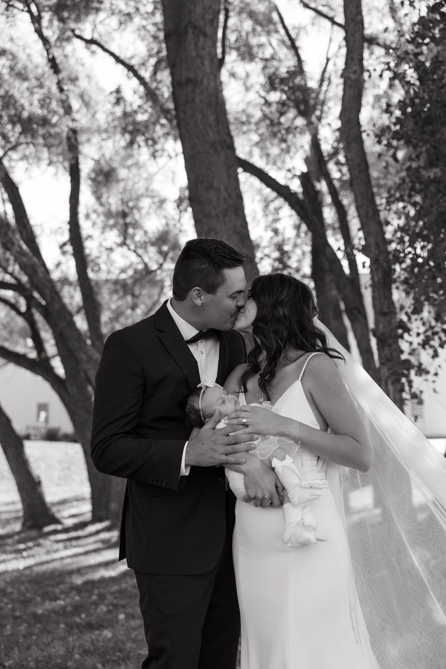 A romantic black and white outdoor wedding portrait with couple embracing under trees.