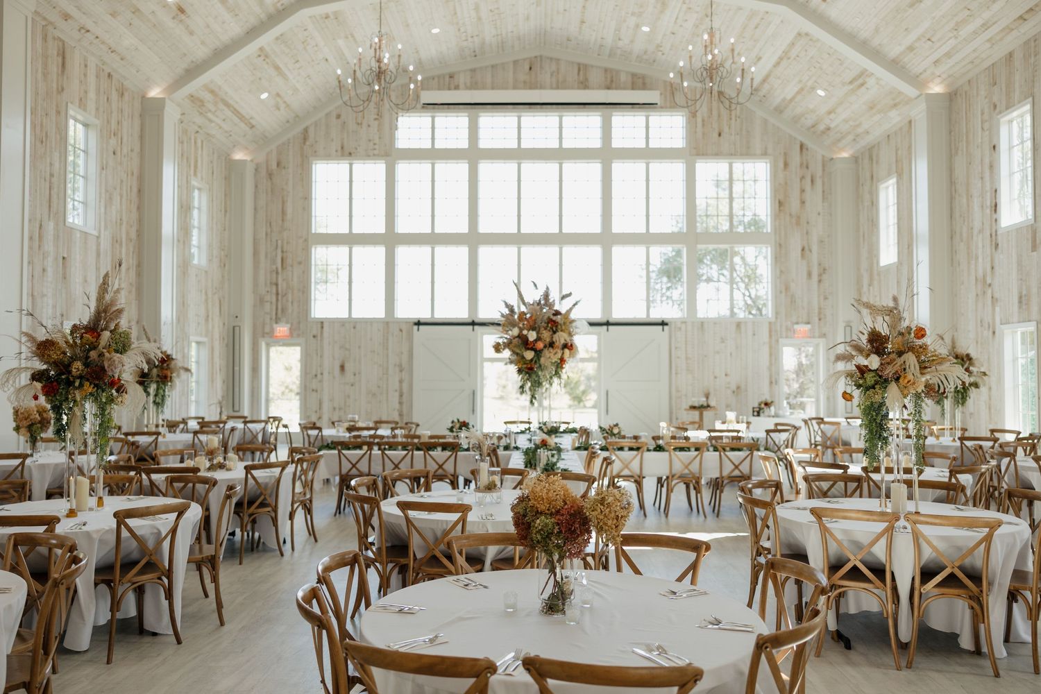Elegant wedding reception venue with white walls, large windows, and rustic wooden tables decorated with floral centerpieces.